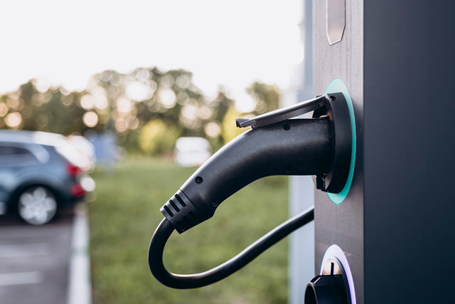 Close-up van een laadpaal voor elektrische voertuigen, met een aangesloten oplaadkabel, geplaatst op een parkeerplaats met groene omgeving op de achtergrond.